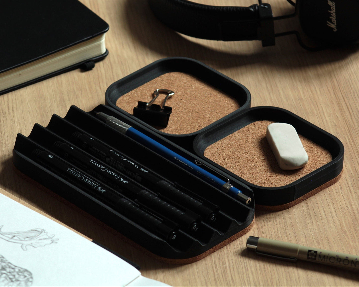 Set of three matte black desk trays with a natural cork bottom, set on a wooden surface. Two small square trays with a cork-lined top and a wavy pen tray with some fineliners on it.
