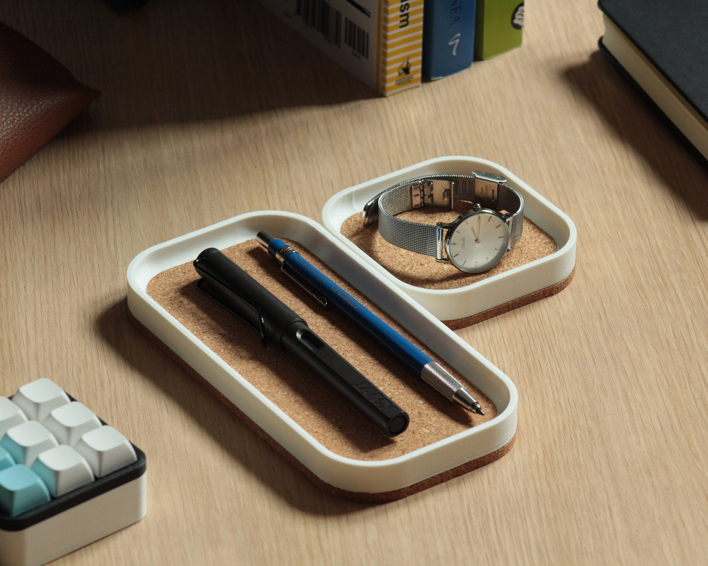 Two matte white magnetic catch-all trays with a natural cork bottom, placed on a wooden desk surface next to a some books and a mechanical macropad.