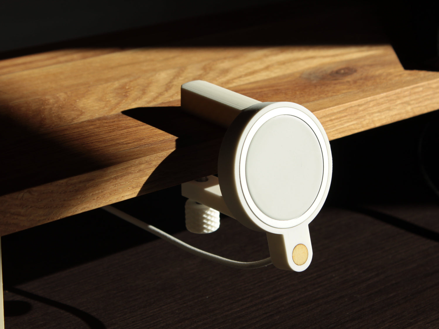 Photo of a minimalistic, matte white magsafe holder clamped to a light oak desk shelf, the sunlight coming from a window on the right side creating a pleasing set of shadows
