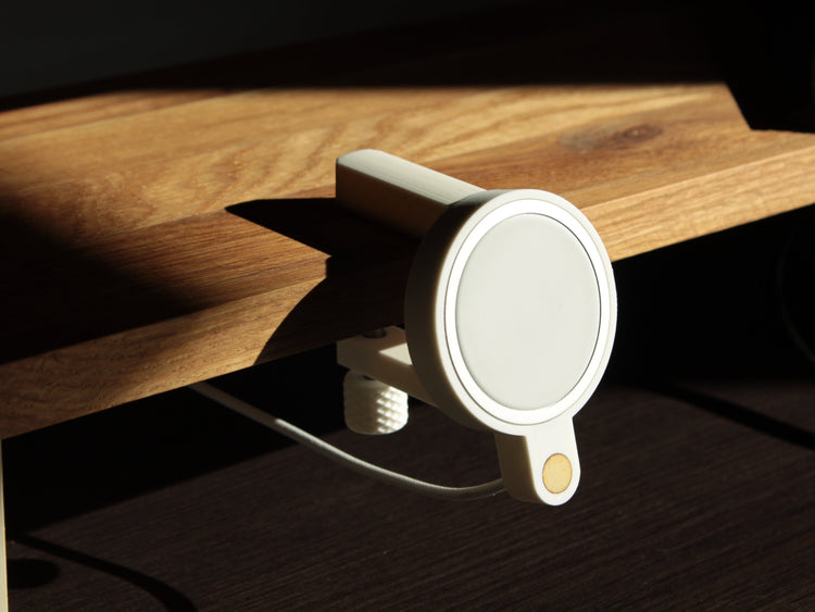 Photo of a minimalistic, matte white magsafe holder clamped to a light oak desk shelf, the sunlight coming from a window on the right side creating a pleasing set of shadows