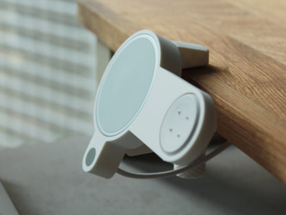 Photo of NODO Lite holder compatible with Google Pixelsnap and Pixel Watch 2 & 3 charger holder clamped on an oak desk shelf. The holder is made of matte white color with a sage green accent dot. Close-up focus on the Pixel Watch 2 & 3 charger with 4 pogo pins