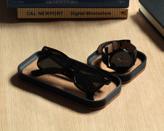 Two black magnetic catch-all trays with a natural cork bottom, placed on a wooden desk surface next to a notebook and a pen.