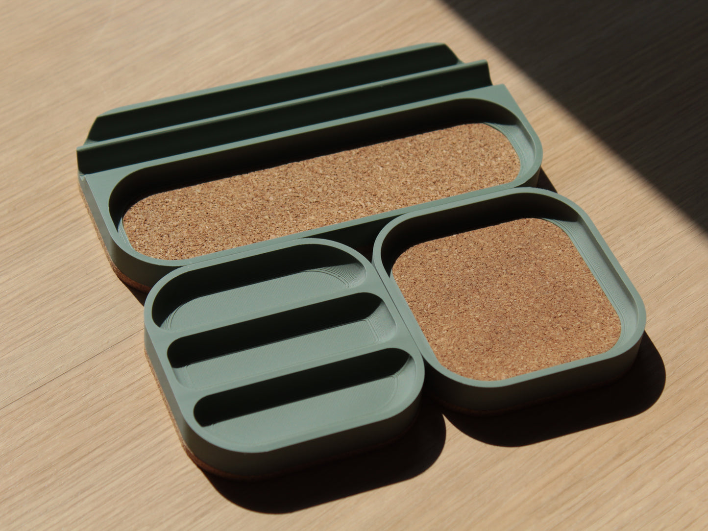 A set of sage green modular trays with rounded corners and cork padding. There are 2 small square trays and a longer one with a cork-padded pocket