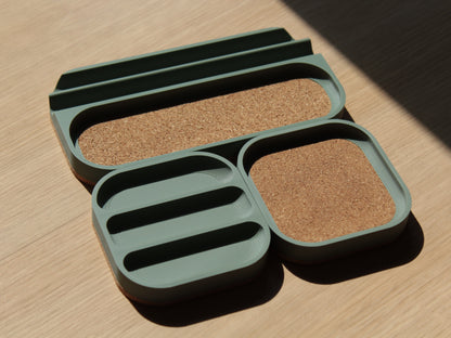 A set of sage green modular trays with rounded corners and cork padding. There are 2 small square trays and a longer one with a cork-padded pocket