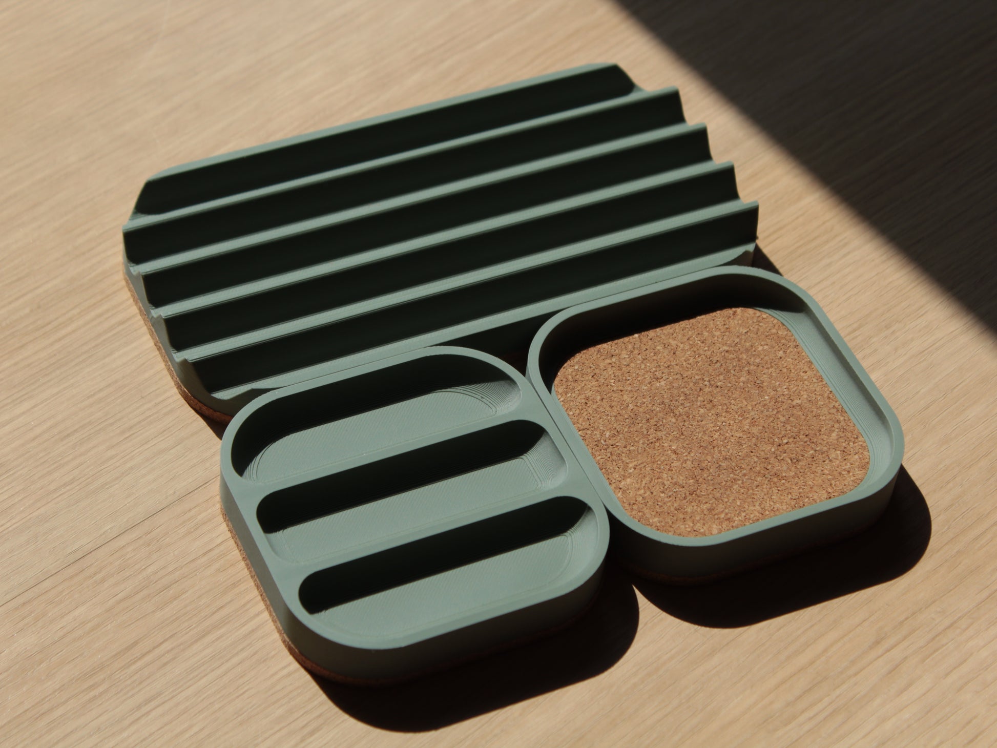 A set of sage green modular trays with rounded corners and cork padding. There are 2 small square trays and a longer one with V slots for holding pens and markers