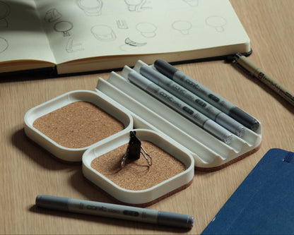 Set of three matte white desk trays with a natural cork bottom, set on a wooden surface. Two small square trays with a cork-lined top and a wavy pen tray with some markers on it.