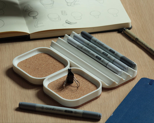 Set of three matte white desk trays with a natural cork bottom, set on a wooden surface. Two small square trays with a cork-lined top and a wavy pen tray with some markers on it.