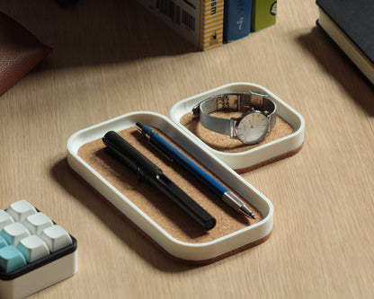 Two matte white magnetic catch-all trays with a natural cork bottom, placed on a wooden desk surface next to a some books and a mechanical macropad.