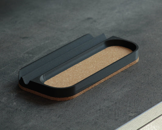 A medium-sized black pen storage tray with a natural cork bottom, placed on a textured surface.