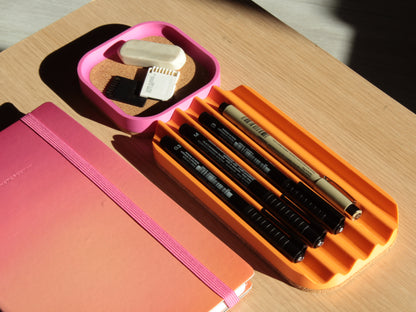 Orange pen tray with pens and a pink notebook on a wooden surface